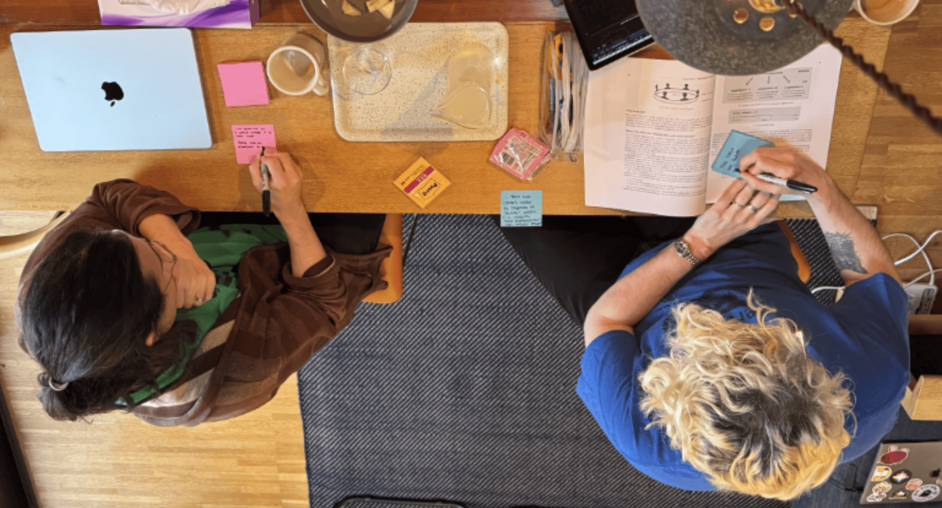 An above-shot photo of two people, one wearing a green t-shirt and another wearing a blue t-shirt, writing on post-it notes and discussing with one another. They are the founders of Studio Terranova, CJ and Matt.)