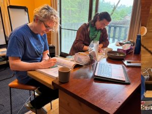 two people working at a desk in a well-lit room. One person is blonde and is wearing a blue shirt. The other is dark-haired and is wearing a green shirt. They look serious as they pore over books and write on sticky notes.